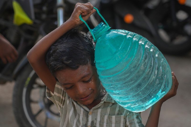 A young boy pours water from a large turquoise plastic container over his head, with motorcycles visible in the background.