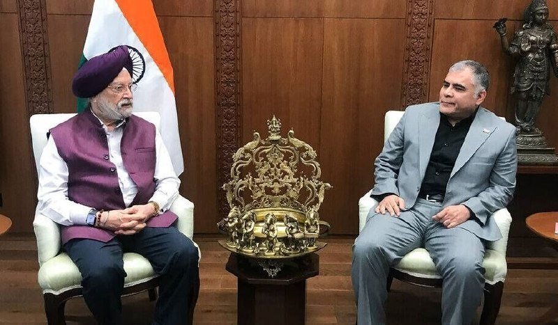 Two men in formal attire sit facing each other in ornate chairs beside an Indian flag, with a golden decorative sculpture between them.