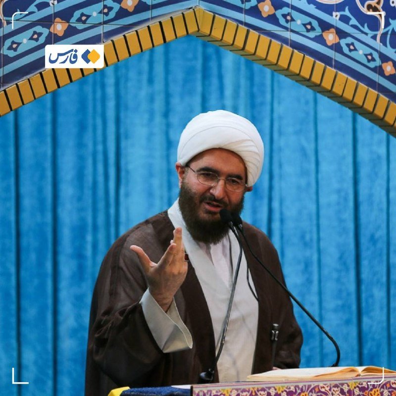 A bearded man in a white turban and glasses speaks into a microphone while gesturing, standing before a blue curtain within a decorated archway.