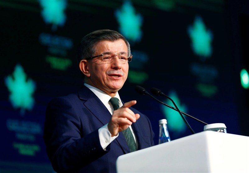 A man in a suit and glasses speaks at a podium with microphones, gesturing with one hand, against a backdrop of green tree logos and text.