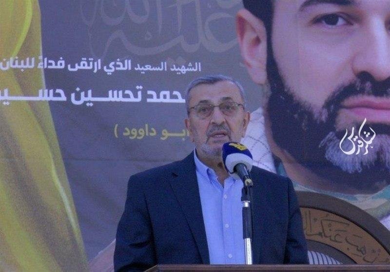 A bespectacled man in a dark blazer and light blue shirt speaks into a microphone, standing before a large poster featuring Arabic text and a portrait of a bearded man.