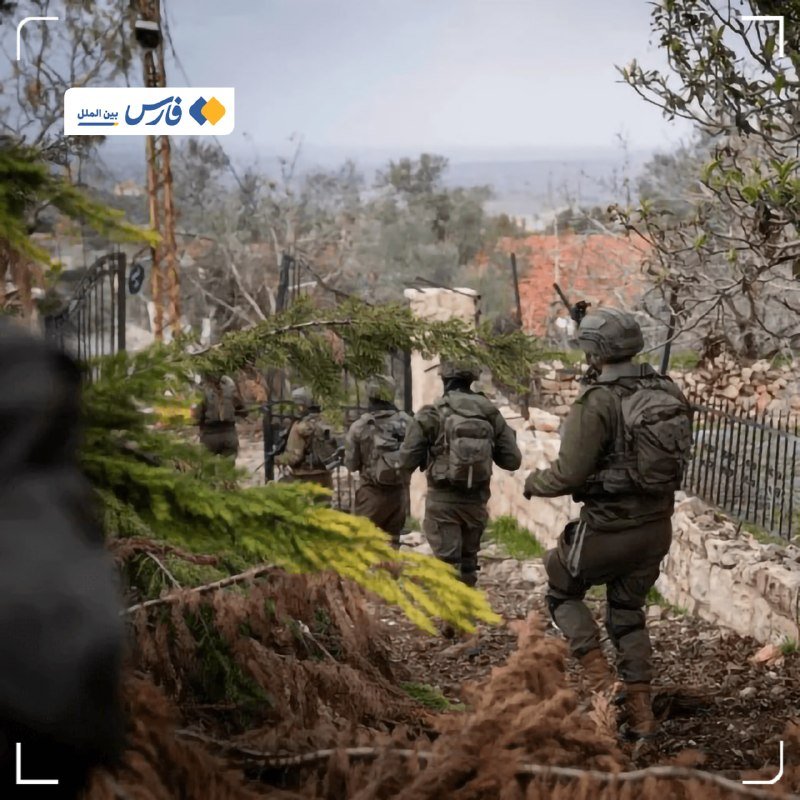 Soldiers in helmets and olive uniforms with backpacks move past rubble and stone walls through a rural, tree-lined area.