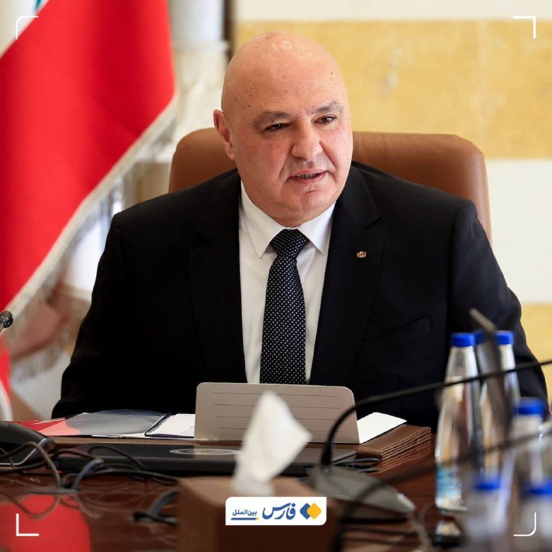 A bald man in a dark suit and polka-dot tie speaks at a desk with documents, a tablet, and water bottles, beside a red-and-white flag.