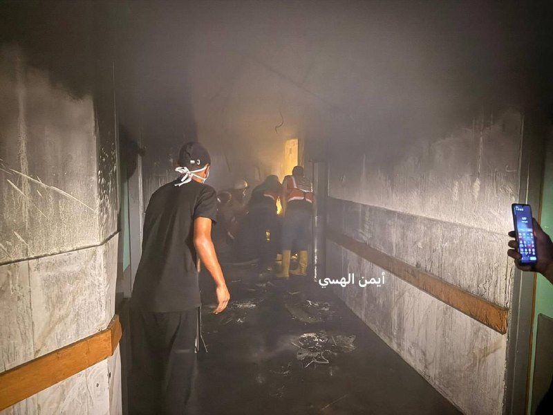 Several people walk through a smoke-filled corridor with charred walls and debris, while a hand in the foreground holds up a smartphone.