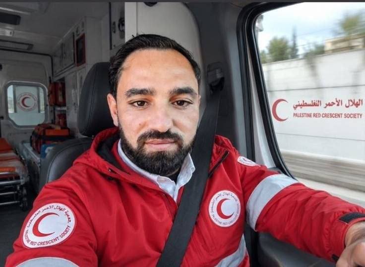 A bearded man wearing a red Palestine Red Crescent Society jacket sits inside a marked ambulance, looking directly at the camera.