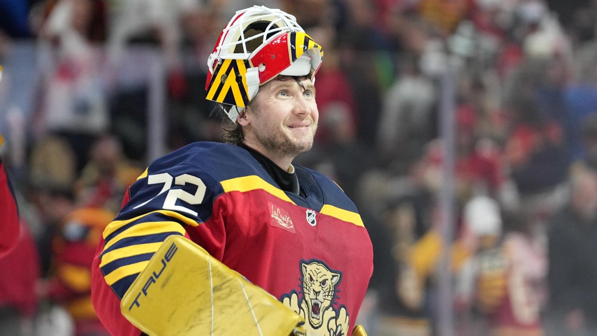A Florida Panthers hockey goaltender wearing jersey number 72 smiles while lifting his red, yellow, and navy goalie mask off his head during a game.