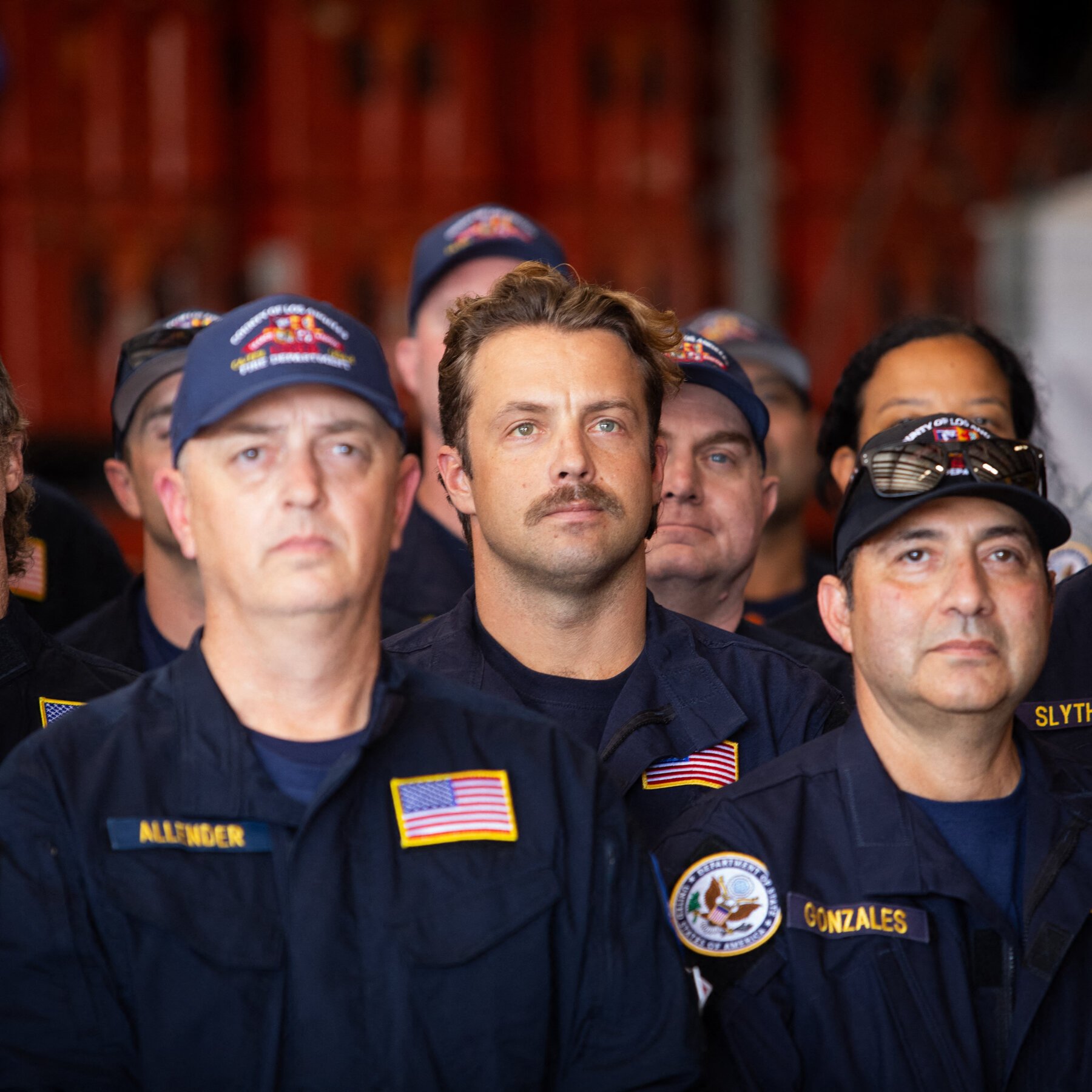 A group of men in dark blue uniforms with American flag patches and "ALLENDER," "GONZALES," and "SLYTH" name tapes stand together, looking upward with serious expressions.