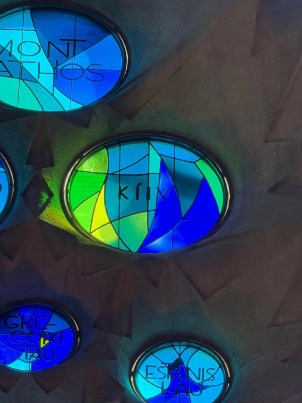 Circular stained glass windows bearing names including "MOUNT ATHOS," "KITH," "GREEN GLYPH," and "ST. ESTBANIS" are set into a stone wall with carved cross patterns.