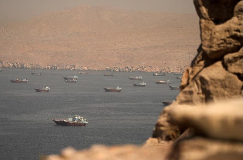 Numerous small boats are anchored in a hazy bay backed by barren, rocky mountains, with blurred rocks framing the foreground.