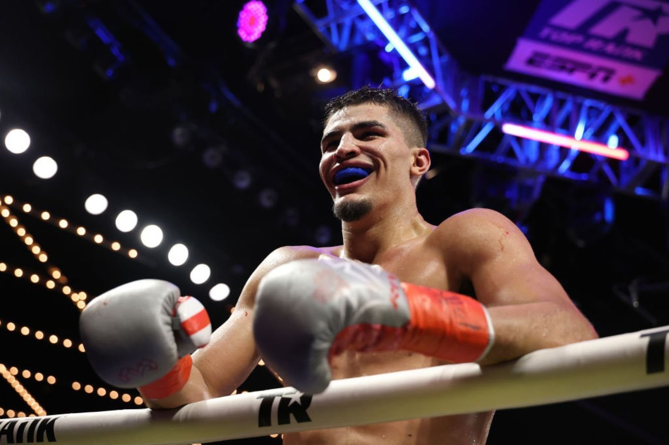 A shirtless boxer wearing a blue mouthguard and red-and-gray gloves smiles while leaning on the ring ropes under bright arena lights.