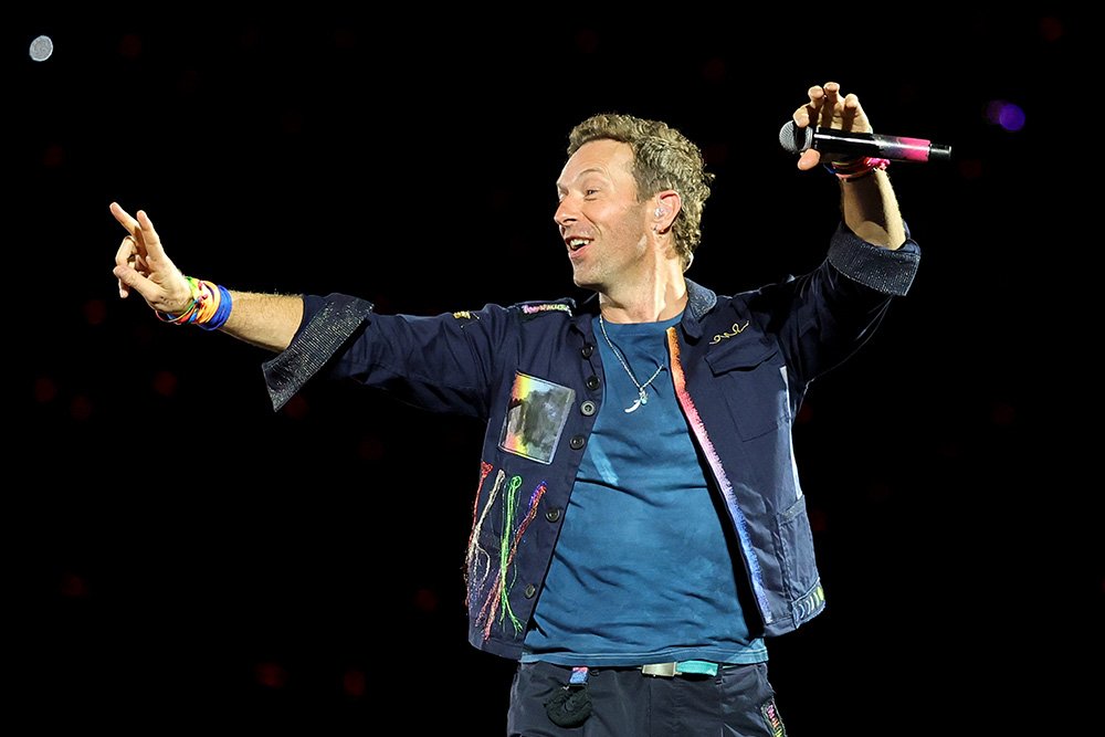 A smiling man with short brown hair, wearing a blue t-shirt and dark embellished jacket, holds a microphone aloft and points while performing on a dark stage.