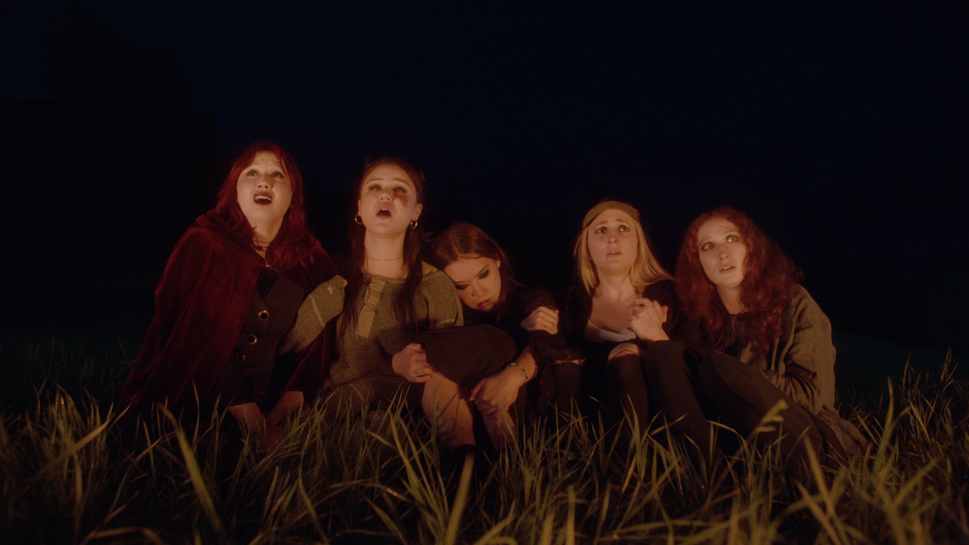 Five young women sit huddled together in tall grass at night, looking upward with expressions of fear and concern.