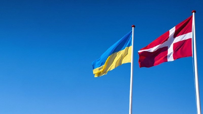 Two flags, one blue-and-yellow and one red with a white cross, wave side by side on flagpoles against a clear blue sky.