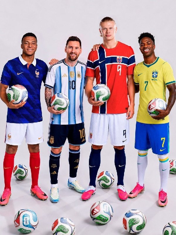 Four soccer players wearing France, Argentina, Norway, and Brazil national team jerseys pose together, each holding a match ball, with additional balls scattered at their feet.