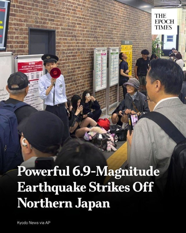 A uniformed official speaks into a red megaphone to people sitting on a station platform beside brick walls, with onlookers filming on phones.