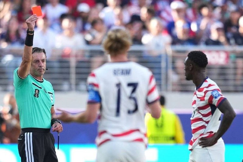 A referee in a green shirt raises a red card above his head toward two soccer players in red, white, and blue striped jerseys during a match.
