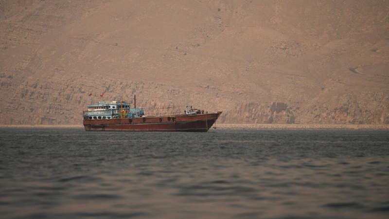 A wooden boat with a white cabin floats on calm water, backed by a massive, barren rocky mountain range under warm light.