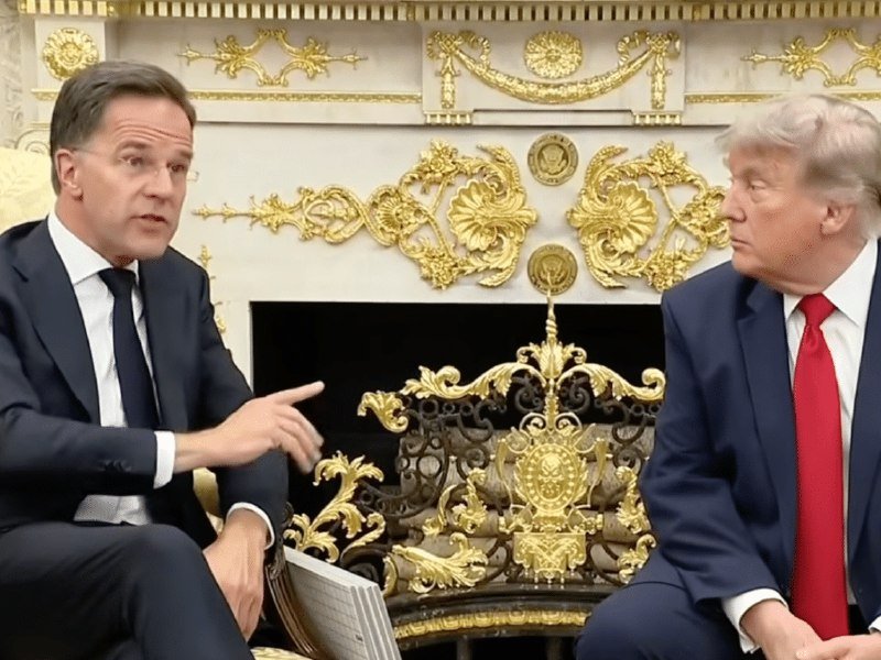 Two men in dark suits sit facing each other in front of an ornate gold-decorated fireplace, one gesturing while speaking.