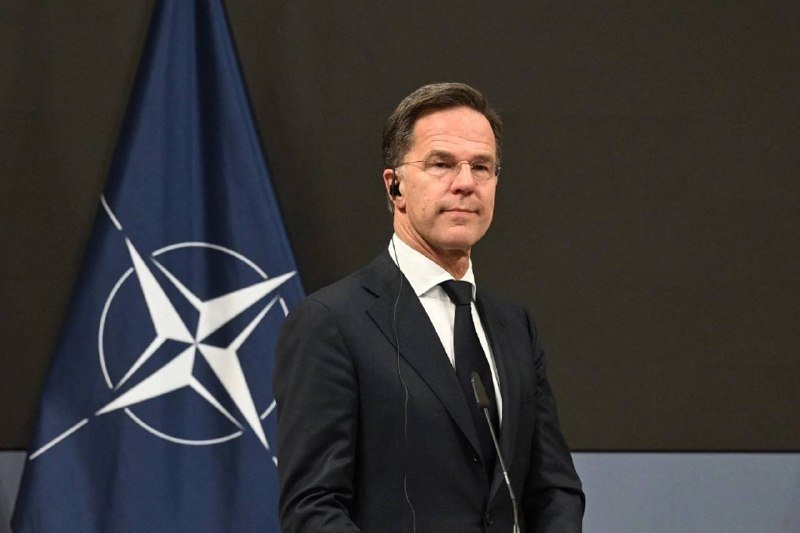 A bespectacled man in a dark suit and tie stands at a microphone beside a NATO flag.
