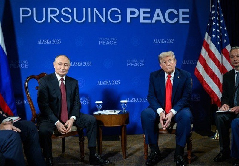 Two suited men sit facing forward in chairs at a table with water glasses, before a blue backdrop reading "PURSUING PEACE" and "ALASKA 2025," with a Russian flag and an American flag visible.