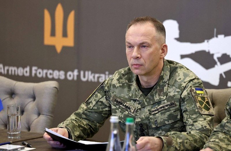 A man in a Ukrainian military camouflage uniform sits at a table holding papers, with a backdrop displaying the Armed Forces of Ukraine emblem and text.