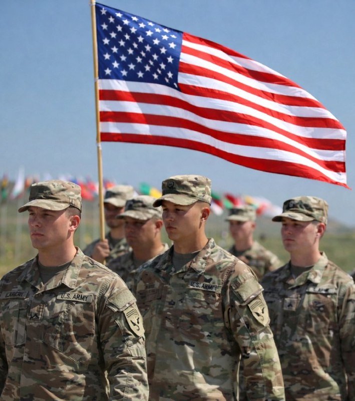 U.S. Army soldiers in camouflage uniforms stand in formation beneath a large American flag, with colorful flags visible in the background.