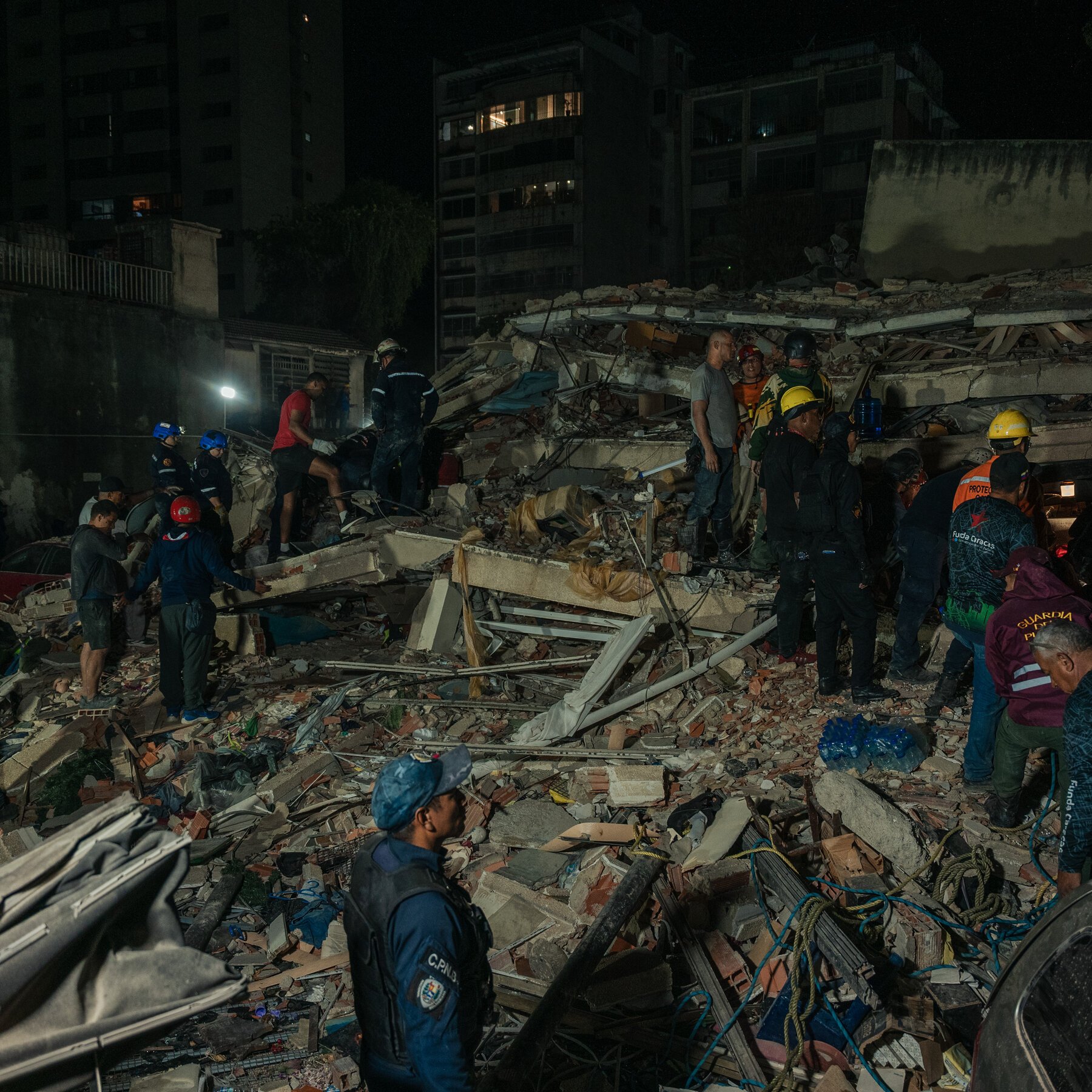 Rescue workers in helmets and uniforms search through a large pile of rubble and collapsed building debris at night.