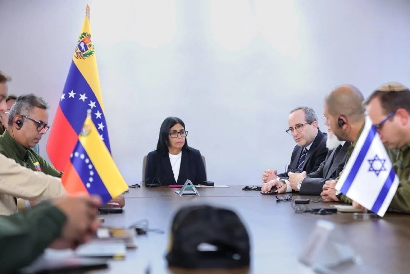 Officials in civilian and military attire sit across a conference table flanked by Venezuelan and Israeli flags.