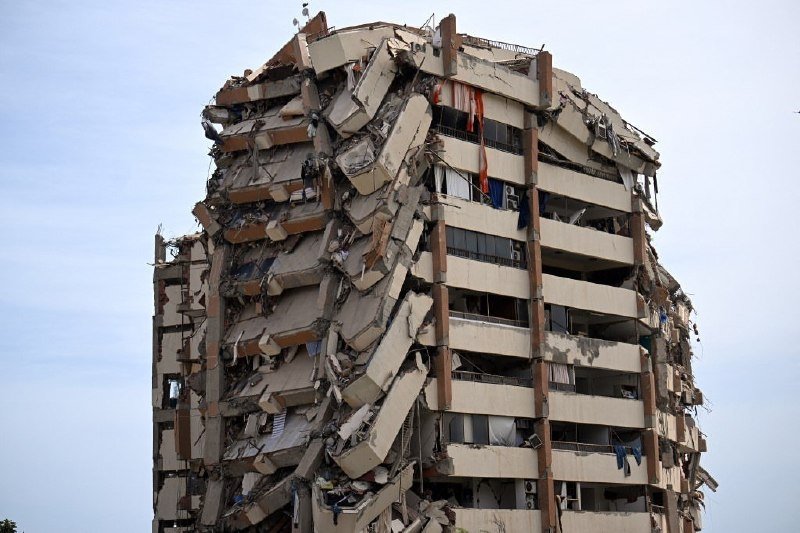 A multi-story building stands partially collapsed, with one side's facade sheared off, exposing twisted concrete slabs and hanging debris against a pale sky.