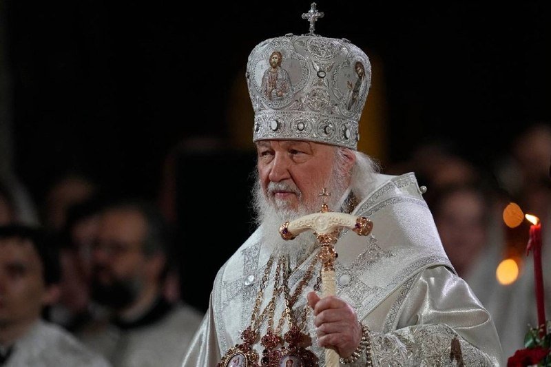 A bearded clergyman wearing a jeweled white mitre and ornate vestments holds a ceremonial staff during a religious service.