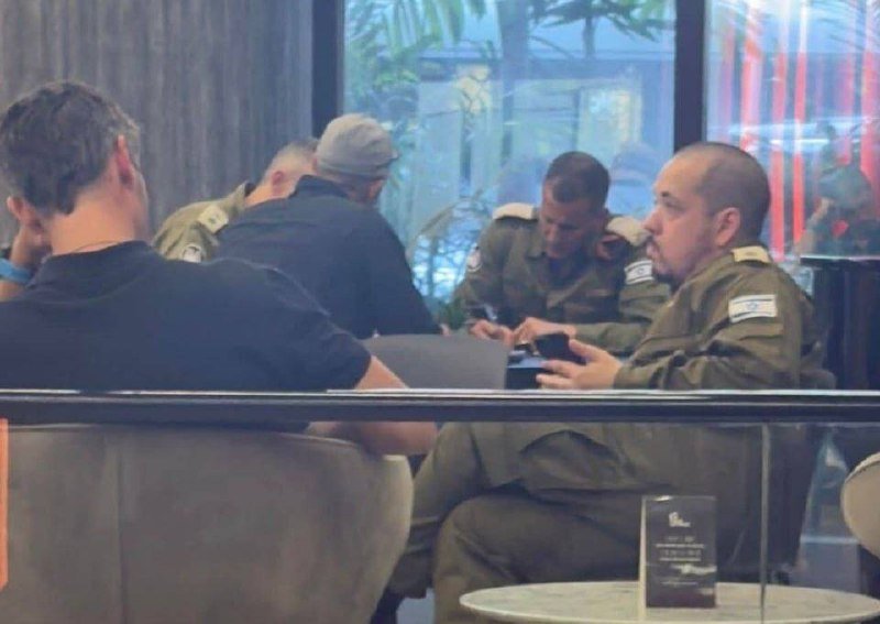 Four men in military uniforms sit at a table in an indoor space with large windows, one looking at his phone.