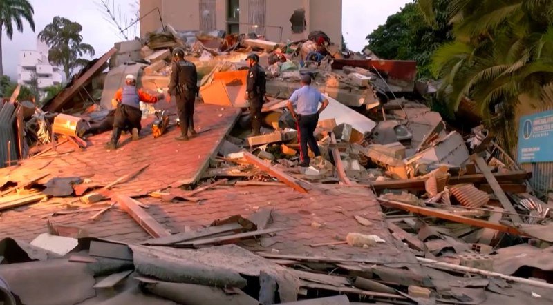 Uniformed personnel and rescue workers stand amid the extensive rubble of a collapsed building, searching through scattered debris and broken tiles at dusk.
