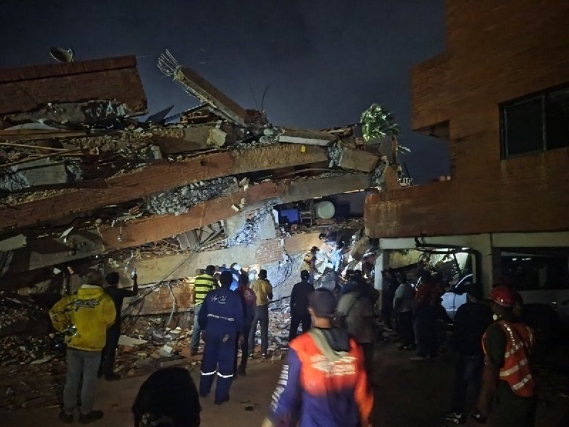 Rescue workers in high-visibility gear gather at night around a collapsed multi-story brick building amid rubble and debris.