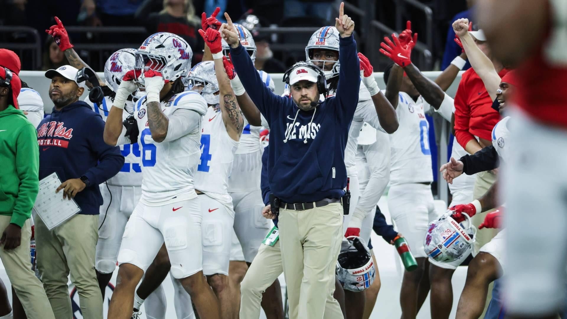 Football players in white uniforms with red gloves and coaches in navy "Ole Miss" hoodies celebrate on the sideline, raising their arms and pointing upward.
