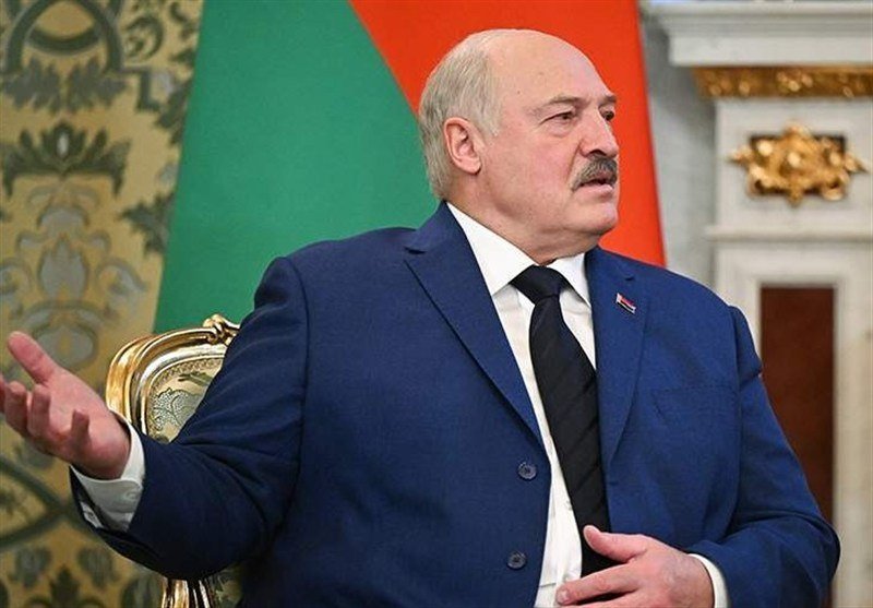 A man with a mustache in a blue suit and black tie gestures with his hand while seated, with a red and green flag visible behind him.