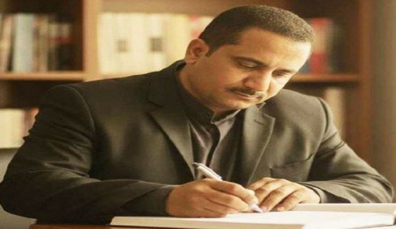 A man in a dark jacket writes in a book at a desk, with bookshelves visible in the background.
