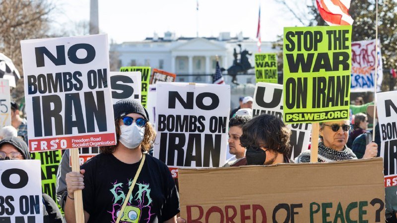 Masked demonstrators hold signs reading "NO BOMBS ON IRAN" and "STOP THE WAR ON IRAN!" in front of a white government building.
