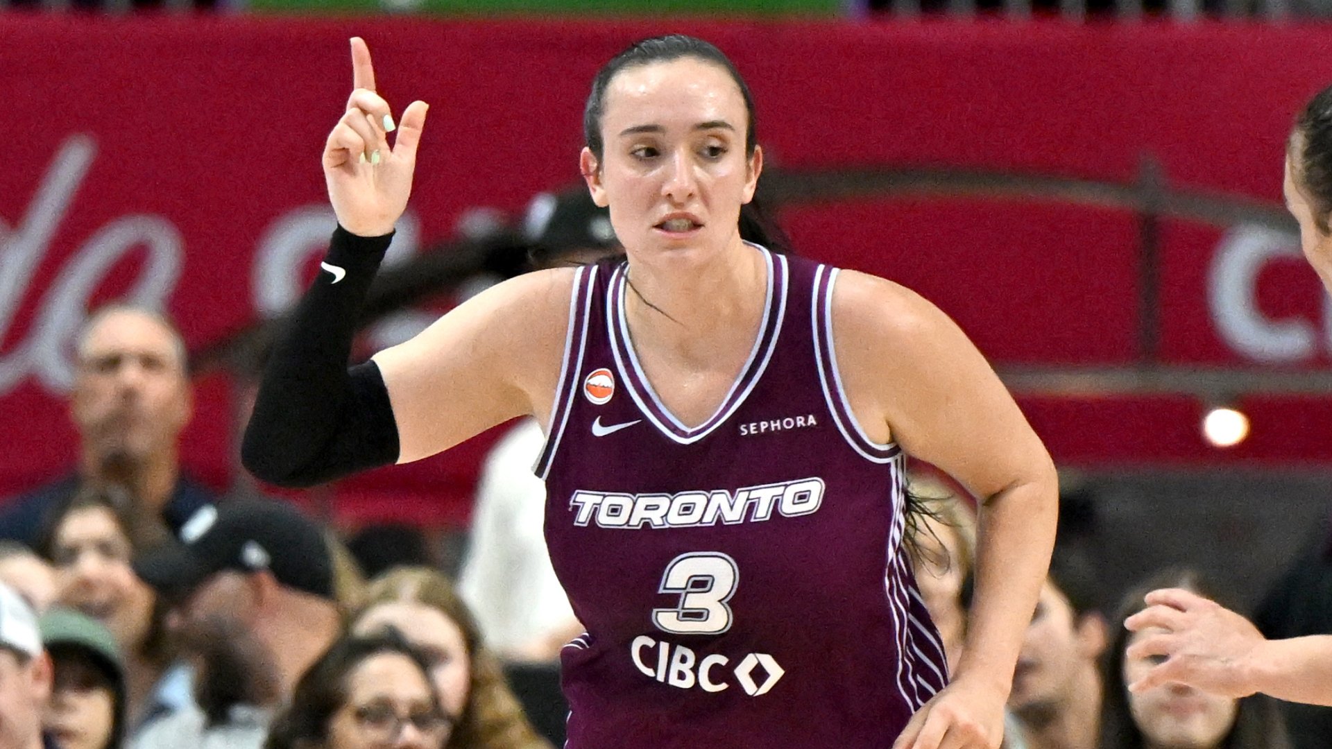 A basketball player wearing a maroon "Toronto" jersey with the number 3 raises her index finger while standing on a court in front of spectators.