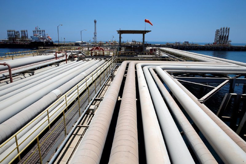 Large industrial pipelines stretch across a coastal loading facility, with port cranes, a tanker ship, and a red windsock visible in the background under a clear sky.