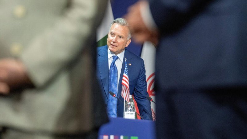 A man in a dark blue suit and tie stands behind a table with a microphone, small American flag, and water bottle, flanked by partially visible people in the foreground.