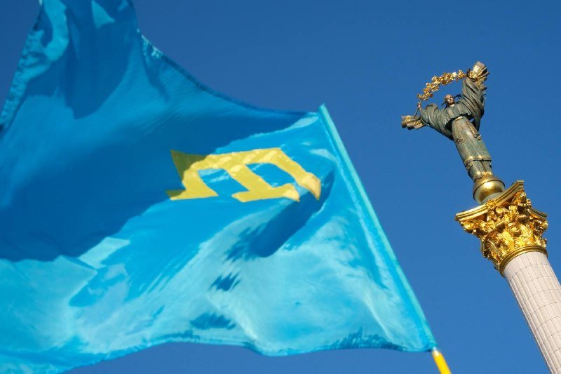 A light blue flag featuring a gold trident-like emblem waves in the wind beside an ornate monument topped with a golden statue against a clear blue sky.