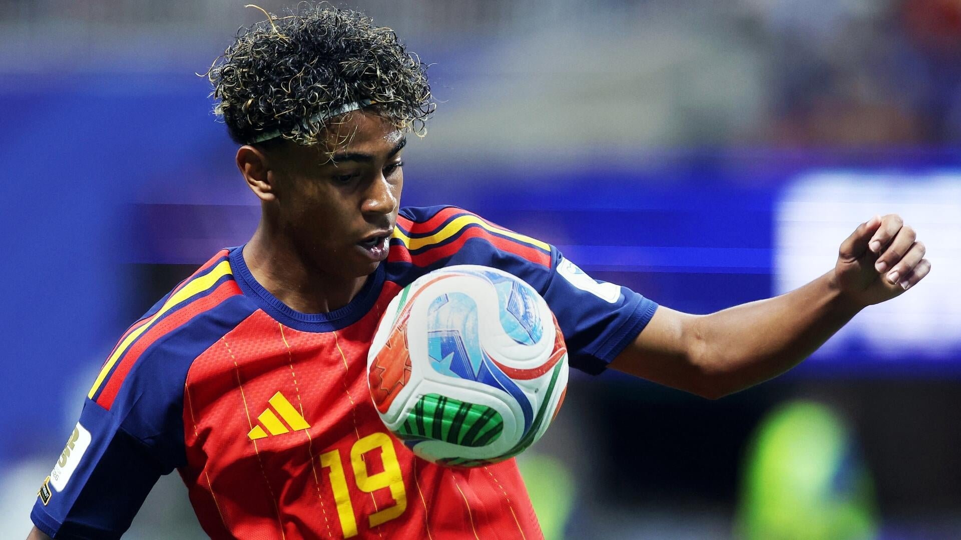 A young soccer player wearing a red and blue jersey with the number 19 controls a colorful match ball during a game.
