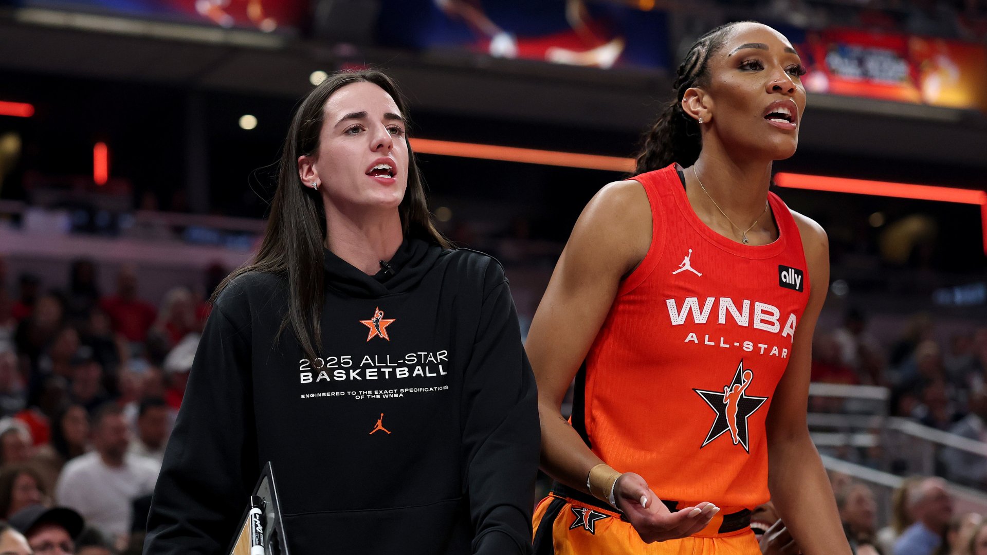 Two women stand courtside at a basketball arena; one wears a black "2025 All-Star Basketball" hoodie, the other a red "WNBA All-Star" jersey, both looking upward.