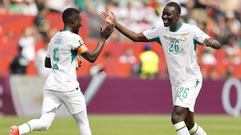 Two soccer players in white and green jerseys, numbers 5 and 26, celebrate with a high-five on a pitch before a crowd.
