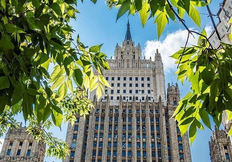 A tall, ornate stone skyscraper with a central spire rises against a blue sky, framed by bright green tree leaves in the foreground.