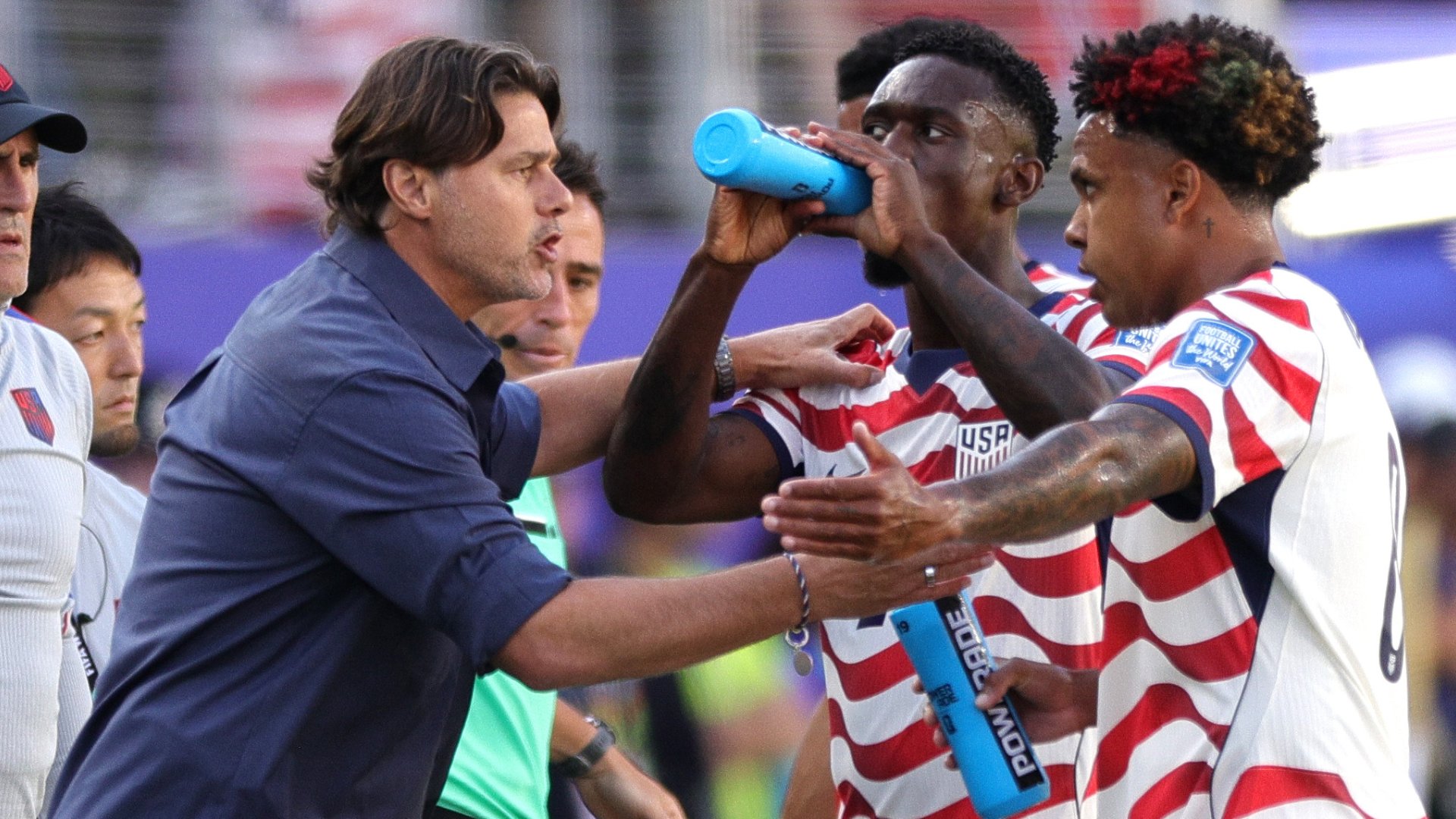 A man in a blue shirt places his hands on a player in a red, white, and blue striped USA jersey who is drinking from a blue water bottle on a soccer field.