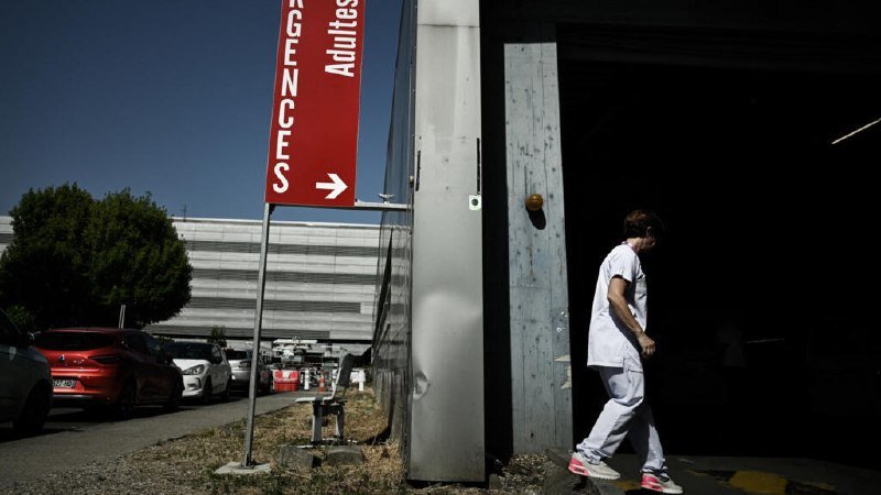 A person in white medical attire walks out of a building entrance near a red sign reading "URGENCES Adultes" with a directional arrow.