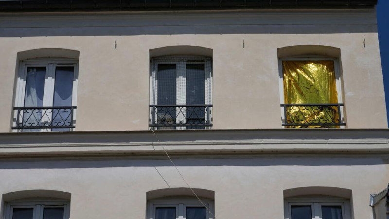 A beige building facade displays three upper windows with wrought-iron balconies, one covered in reflective gold foil, alongside two lower windows beneath a tiled roofline.