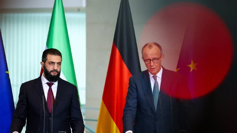 Two men in dark suits stand at podiums during a press conference, with EU, Syrian, German, and Chinese flags displayed behind them.