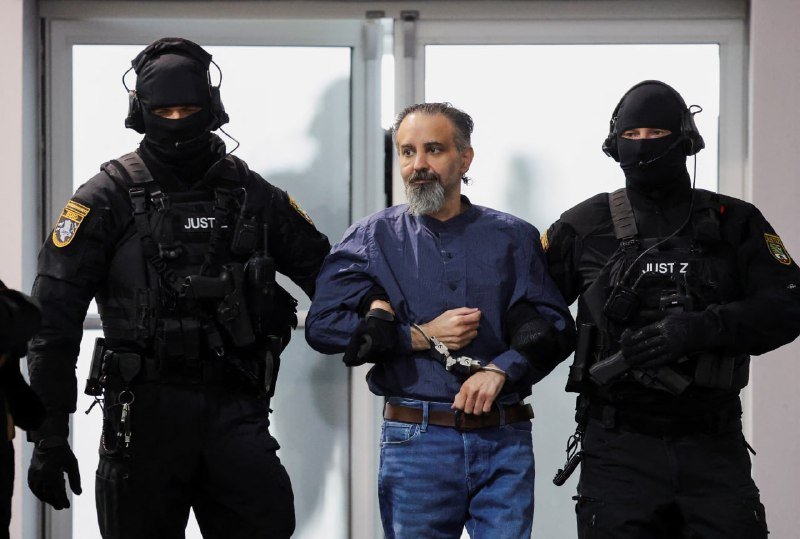 Two masked officers in black tactical gear marked "JUSTIZ" escort a handcuffed, bearded man in a blue shirt and jeans through a glass doorway.